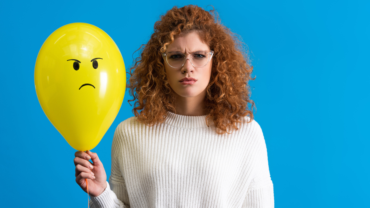 Aggressive woman holding yellow balloon with angry face, isolated on blue