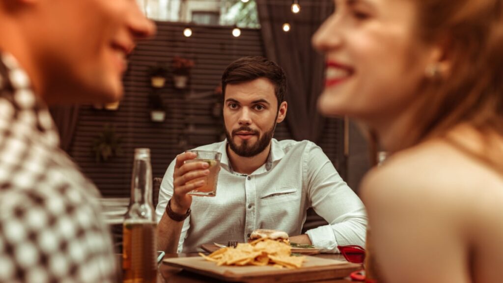 Staring at friends. Attractive nice-looking appealing calm handsome man with short dark hair and beard holding a drink in a hand and staring at his flirting friends at the bar table.