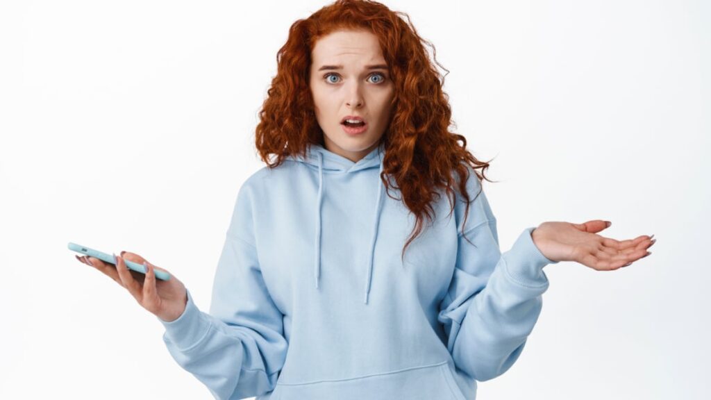 Confused and questioned redhead woman shrugging shoulders while holding mobile phone.