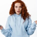 Confused and questioned redhead woman shrugging shoulders while holding mobile phone.