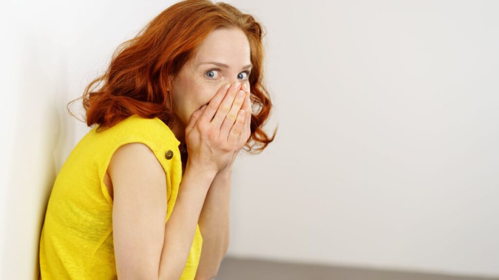 Guilty or embarrassed young redhead woman covering her lower face with her hands and glancing sideways at the camera with big eyes as she leans against a wall.