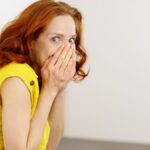 Guilty or embarrassed young redhead woman covering her lower face with her hands and glancing sideways at the camera with big eyes as she leans against a wall.