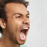 Half profile of aggressive male with dark curly hair, opens mouth widely, screams in panic,