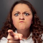 angry young woman pointing at camera over grey background.