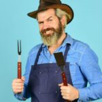 Bbq food. Tools roasting meat. Man in apron hold barbecue grill. Hipster dyed beard promoting bbq equipment.