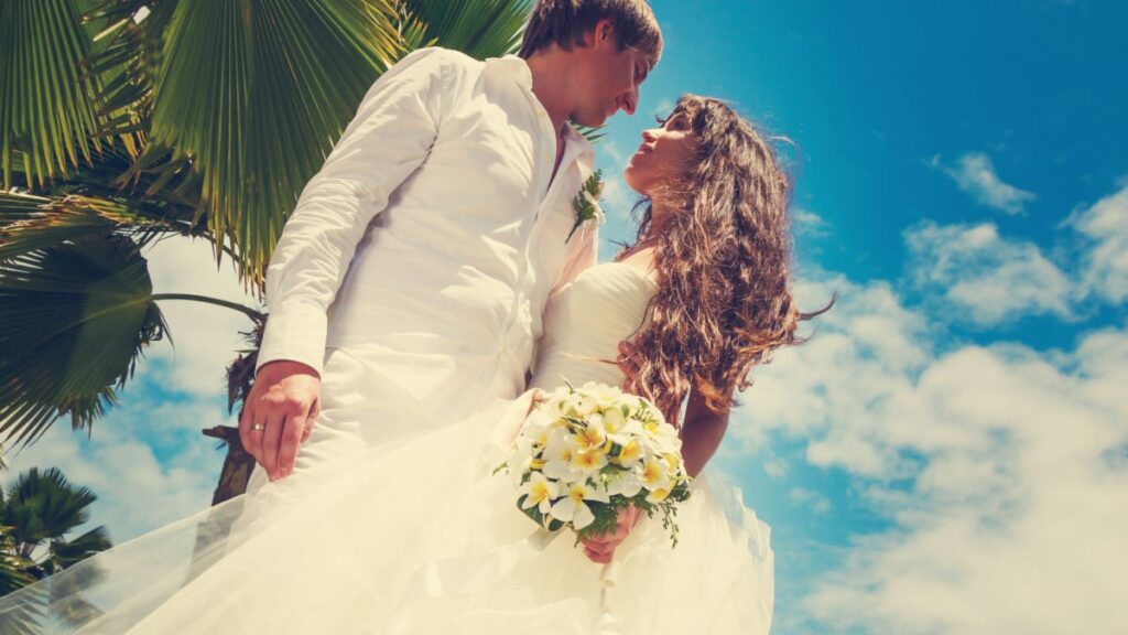 groom and bride in white with a wedding bouquet with frangipani flowers on a background of palm trees and blue sky in the Seychelles.