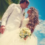 groom and bride in white with a wedding bouquet with frangipani flowers on a background of palm trees and blue sky in the Seychelles.
