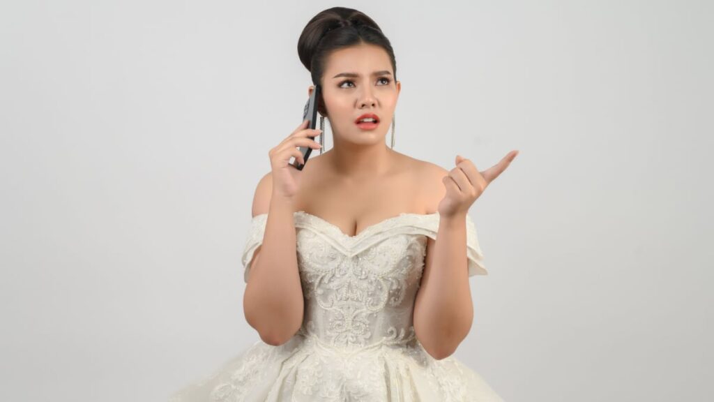Portrait Young asian beautiful bride grimace in white wedding dress holding smartphone in hand and point finger on white background.