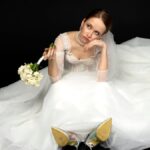 A beautiful charming girl, the bride in a luxurious and elegant white wedding dress, sadly and upset sitting on a floor with a graceful bouquet of white roses. Isolated on a black background.