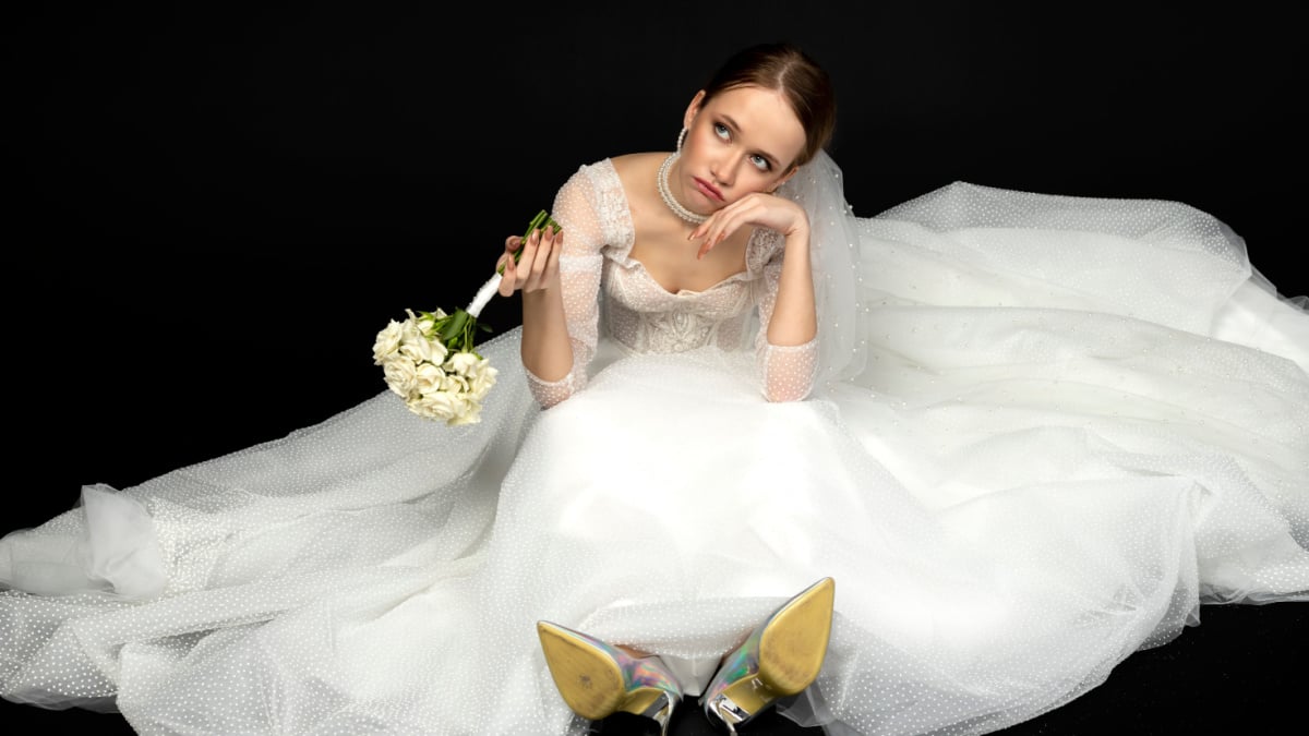 A beautiful charming girl, the bride in a luxurious and elegant white wedding dress, sadly and upset sitting on a floor with a graceful bouquet of white roses. Isolated on a black background.