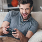 Excited young handsome man playing video game and keeping mouth open while sitting on the couch at home.