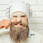 Angry handsome man, cook or baker with flour on face and beard in chef uniform and hat pulls hair from moustache on white brick wall.