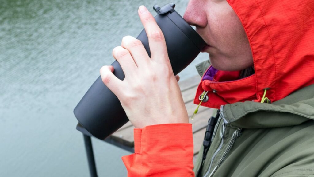 a young frozen girl drinks hot tea from a thermos on the shore of a cold lake.