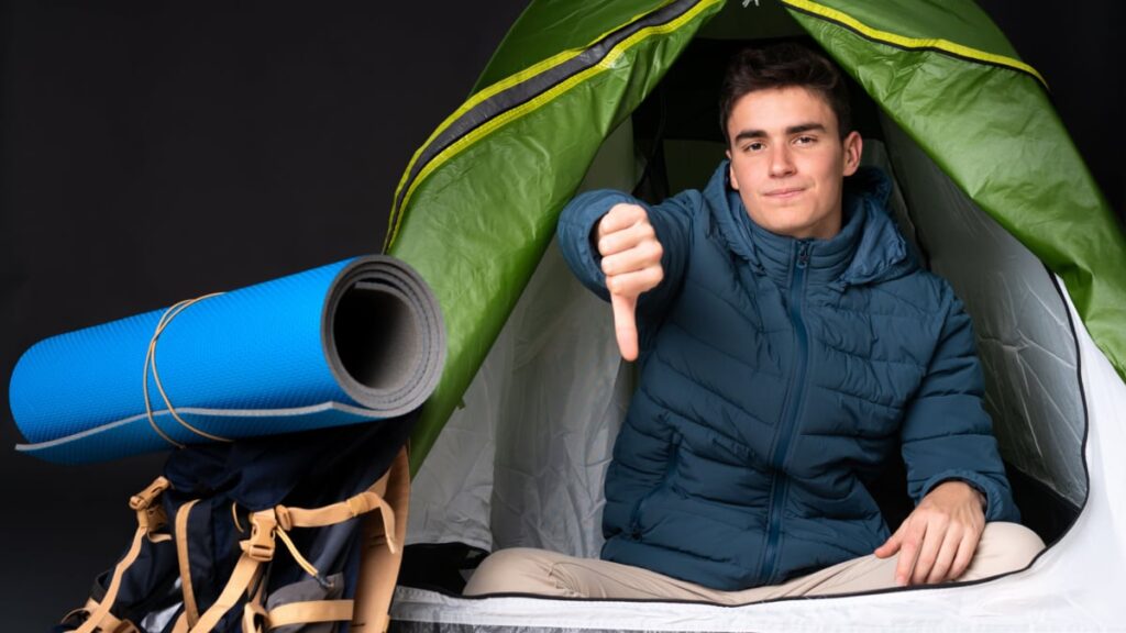 Teenager caucasian man inside a camping green tent isolated on black background showing thumb down with negative expression.