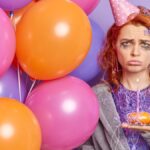 Frustrated unhappy redhead Caucasian woman with spoiled makeup sad to be lonely on her birthday holds cupcake with burning candle bunch of inflated balloons has party at home wears cone hat on head.