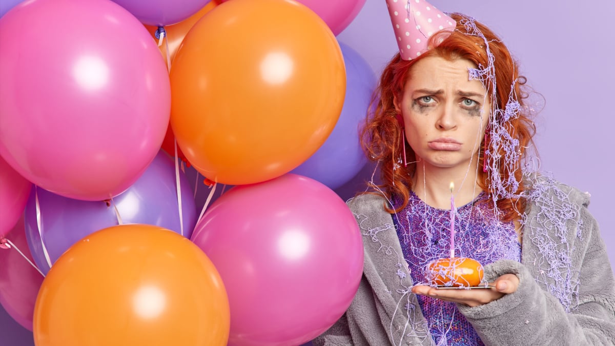 Frustrated unhappy redhead Caucasian woman with spoiled makeup sad to be lonely on her birthday holds cupcake with burning candle bunch of inflated balloons has party at home wears cone hat on head.
