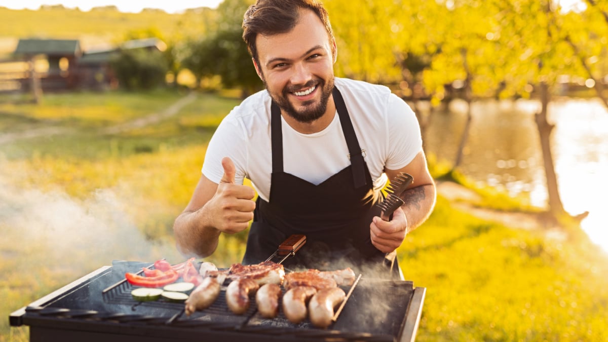 Delighted young ethnic bearded man in black apron smiling at camera and showing thumb up gesture, while preparing delicious grilled sausages and vegetables during picnic in park at sunset.
