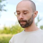 sad guy, young charismatic man in beautiful gold-rimmed glasses, beige t-shirt tiredly closed eyes, green garden in background, concept of eye illness, poor vision, relieve stress in nature.