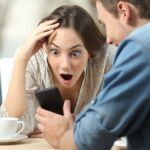 Surprised couple meeting in a coffee shop watching media content in a smart phone.